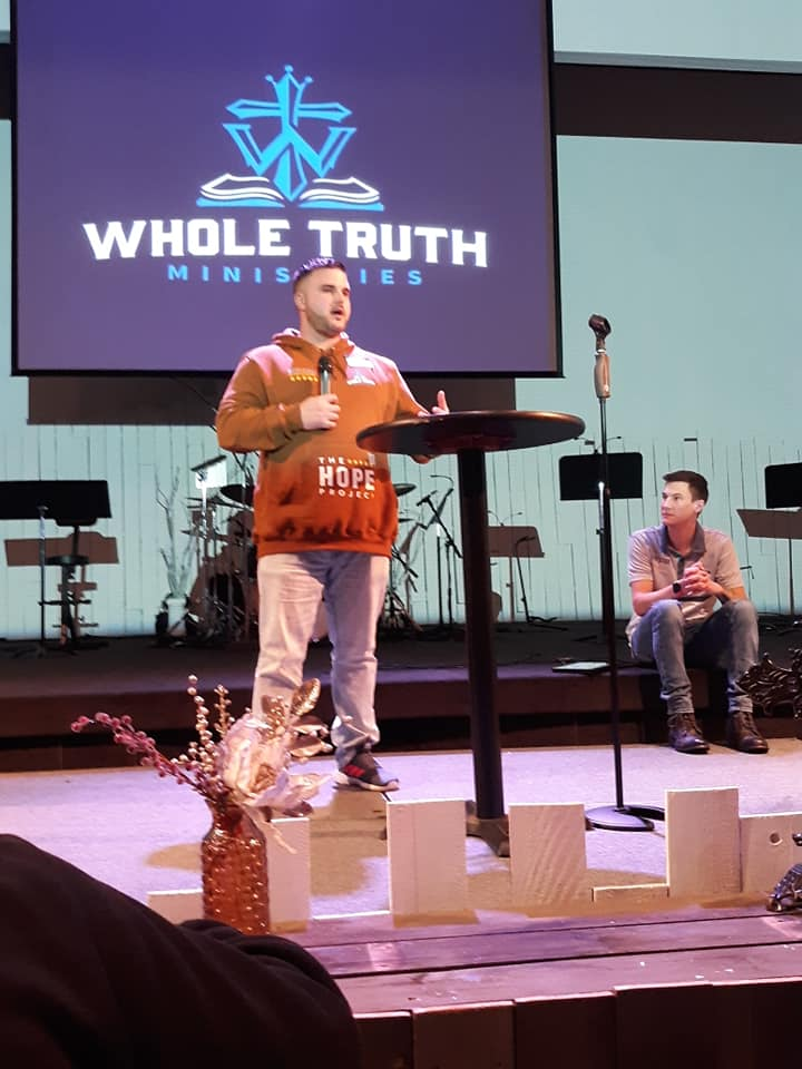 Speaker at Whole Truth Ministries giving a message on stage with the ministry logo displayed on the screen behind him.